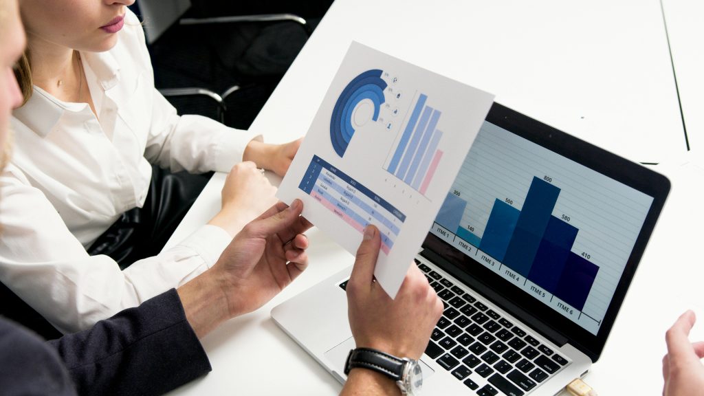 Zwei Personen analysieren Diagramme an Laptop und auf Papier.
