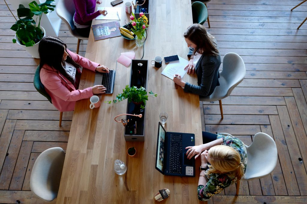 Draufsicht auf einen Holztisch in einem modernen Co-Working-Space, an dem vier Personen mit Laptops und Notizbüchern arbeiten.