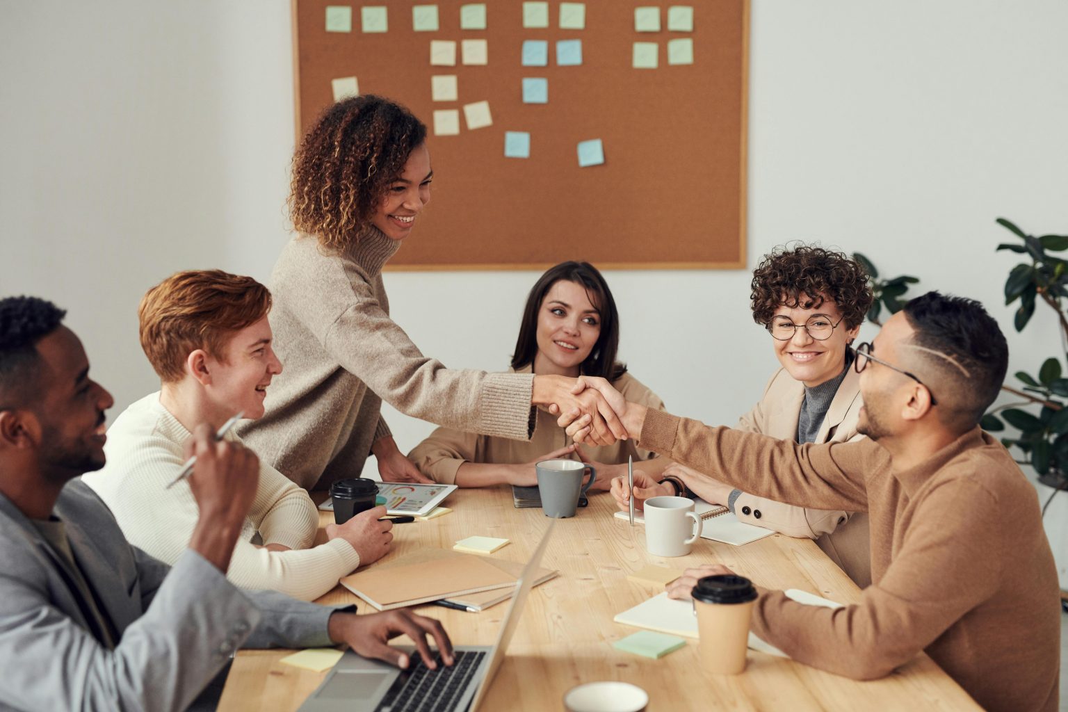 Hintergrundbild. Business-Meeting im modernen Büro: Sechs Kollegen am Konferenztisch, zwei geben sich die Hand, umgeben von Laptops, Notizbüchern und Kaffeetassen. Kreative Teamarbeit vor Pinnwand mit bunten Haftnotizen.