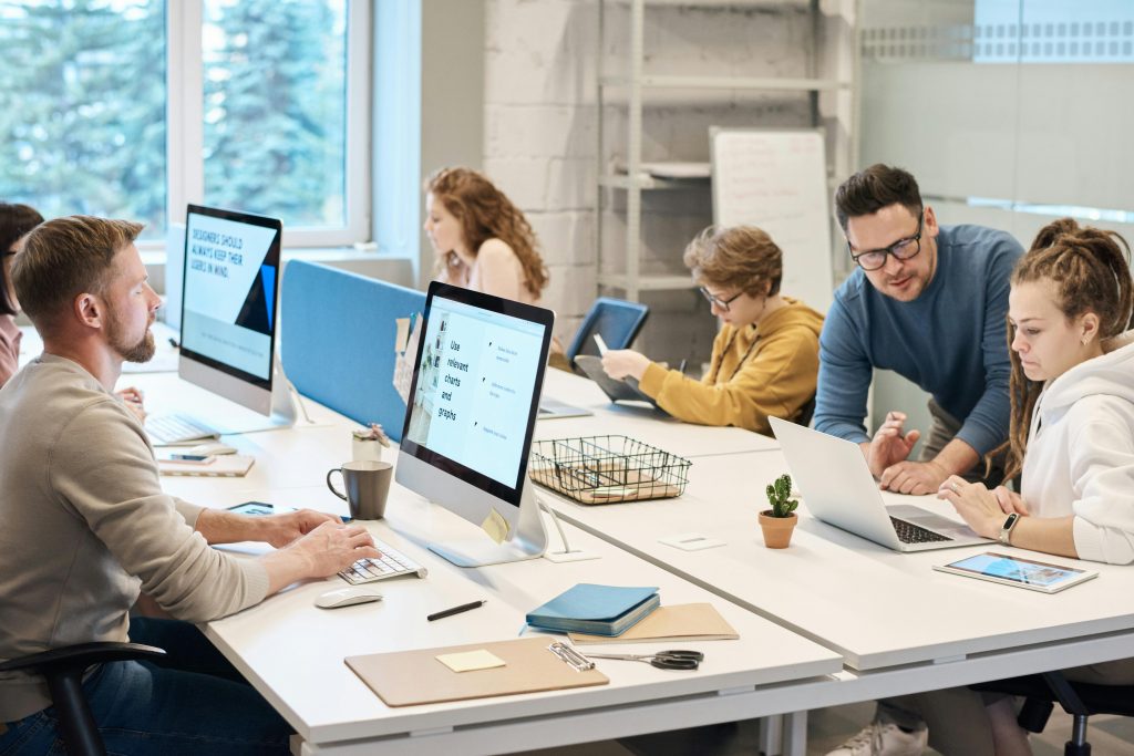 Sechs Personen arbeiten gemeinsam in einem modernen Büroraum. Jede Person nutzt einen Computer. Im Hintergrund ist ein Whiteboard.