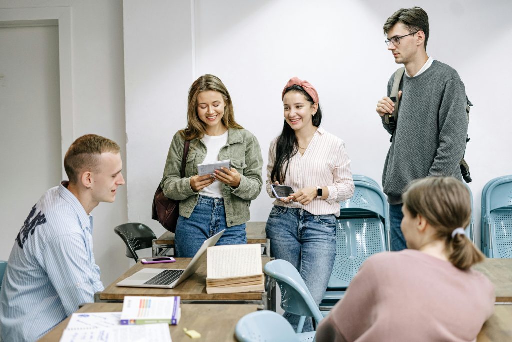Fünf Personen in einem Lern-Setting, vier davon stehen und unterhalten sich, während eine Person an einem Schreibtisch mit aufgeklapptem Laptop und Notizmaterial sitzt.
