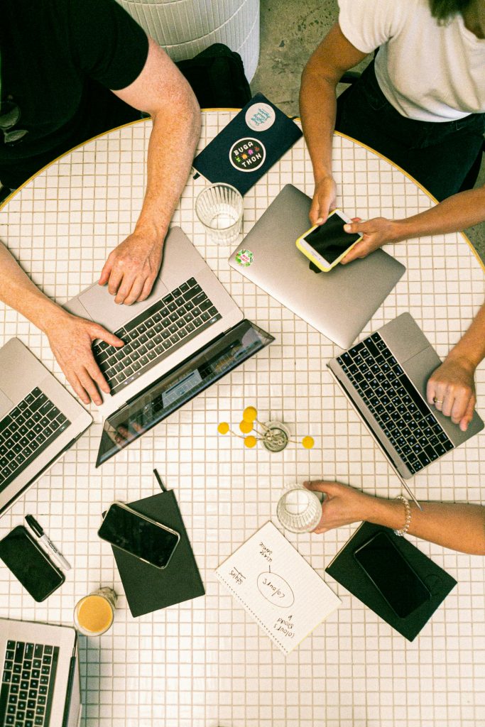 Draufsicht auf einen runden Tisch, an dem vier Personen mit Laptops arbeiten. Auf dem Tisch liegen Smartphones, Notizbücher und Laptops.