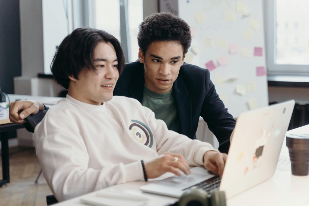 Zwei Personen sitzen an einem Schreibtisch und arbeiten gemeinsam an einem Laptop. Die eine Person zeigt der anderen etwas.