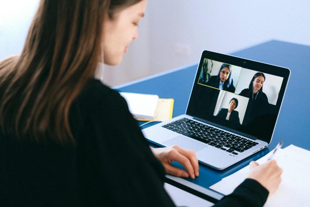Eine Person sitzt an einem Schreibtisch und nimmt an einer Videokonferenz über einen Laptop teil. Auf dem Bildschirm sind vier weitere Teilnehmende zu sehen. Auf dem Schreibtisch liegen ein Stift, Papier und Notizbücher.